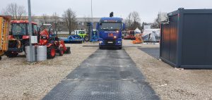 Na obrazie widoczny jest niebieski autobus na placu ze sprzętem budowlanym, wokół szerości i kontenerów.