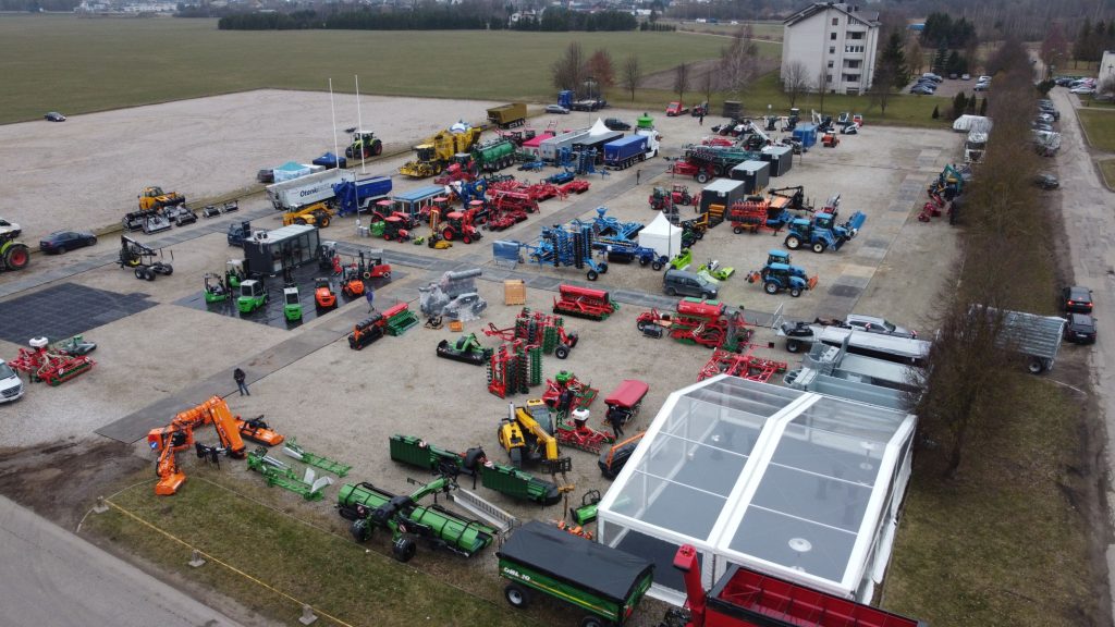 Teren targowy z różnorodnymi maszynami rolniczymi i sprzętem, w otoczeniu pól i budynków.