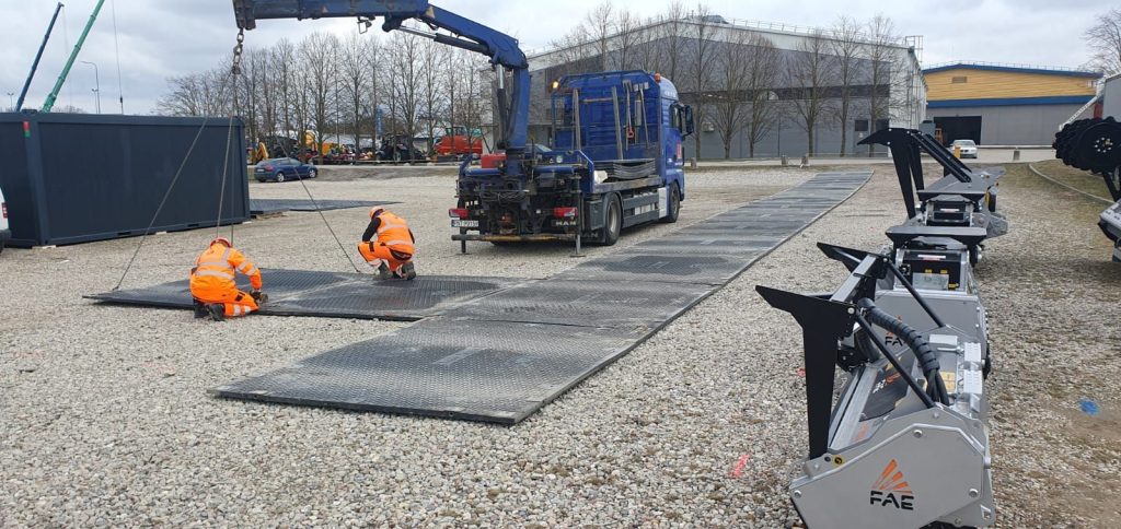 Na obrazie widać dwóch pracowników w pomarańczowych kombinezonach, którzy montują metalowe rampy obok ciężarówki. Obok znajdu