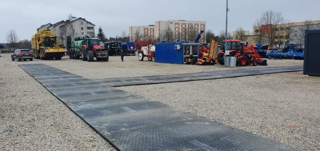 Teren wystawowy z maszynami rolniczymi, przyczepami i kontenerami, w tle budynki mieszkalne. Szare niebo.