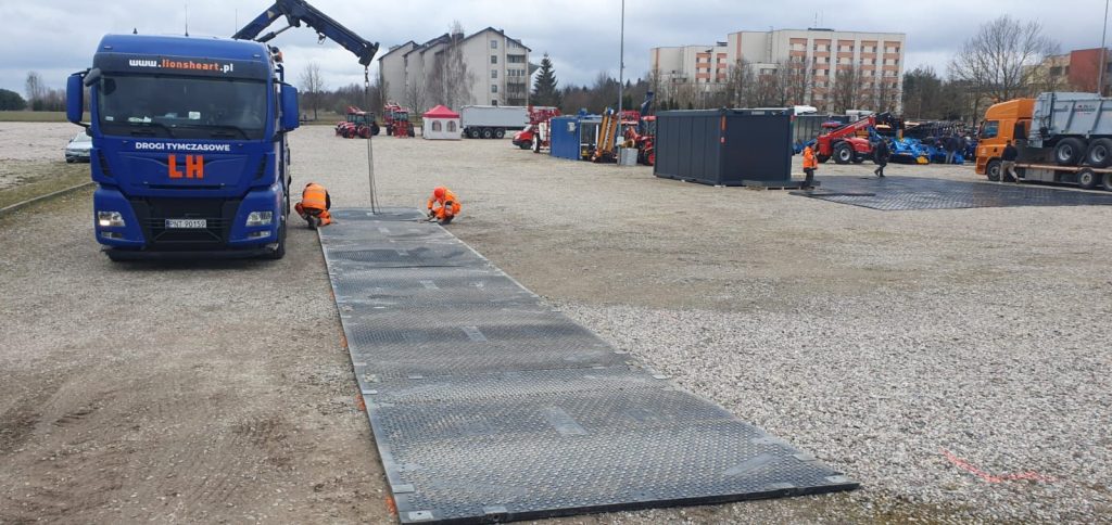 Ciężarówka na placu, dwie osoby w pomarańczowych strojach montują rampę, w tle widoczna flota sprzętu budowlanego.