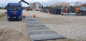 Ciężarówka na placu, dwie osoby w pomarańczowych strojach montują rampę, w tle widoczna flota sprzętu budowlanego.