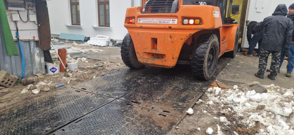 Pomarańczowy wózek widłowy stoi na metalowej platformie, obok leży śnieg i gruz, w tle widać budynek.