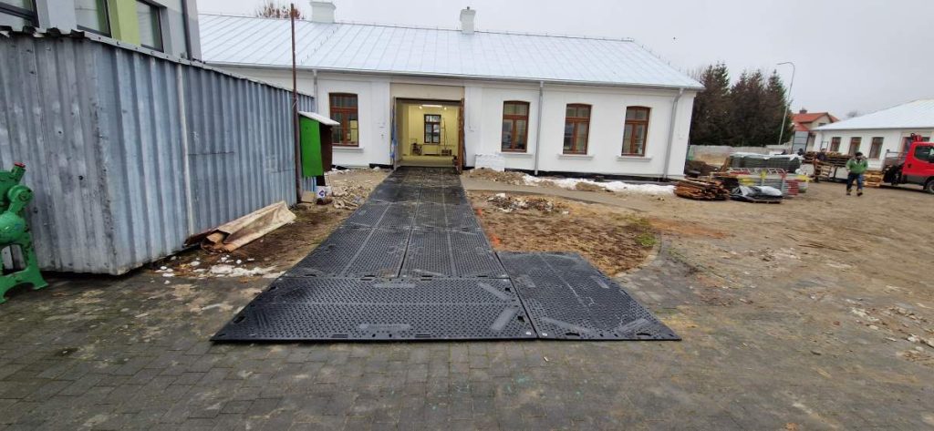 Czarna nawierzchnia prowadząca do budynku, otoczona placem budowy i sprzętem, w tle widoczny biały budynek.