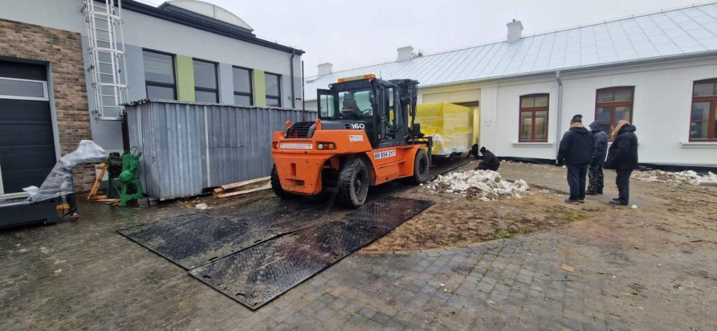 Na zdjęciu widać pomarańczowy wózek widłowy na zewnątrz budynku, obok dwóch osób oraz metalową kontenerową konstrukcję.