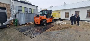 Na zdjęciu widać pomarańczowy wózek widłowy na zewnątrz budynku, obok dwóch osób oraz metalową kontenerową konstrukcję.