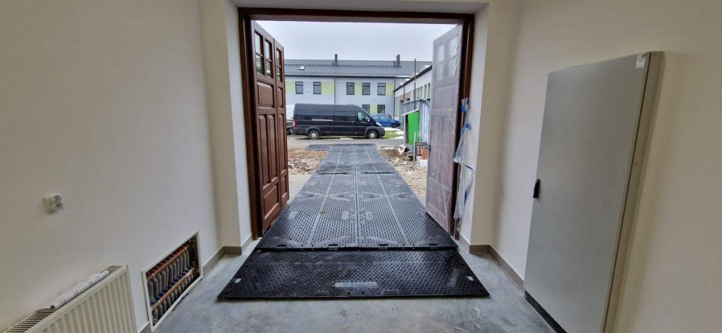 Widok z wnętrza budynku na otwarte drzwi, za którymi znajduje się podjazd i pojazd zaparkowany na zewnątrz.