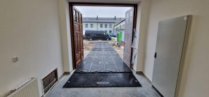 Widok z wnętrza budynku na otwarte drzwi, za którymi znajduje się podjazd i pojazd zaparkowany na zewnątrz.