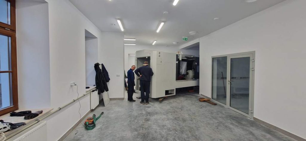 Dwaj mężczyźni stoją przy maszynie w nowoczesnym, jasnym pomieszczeniu. Na podłodze widoczny beton.