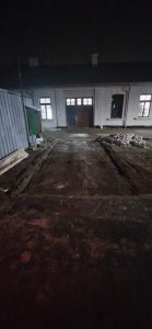 Widok na wyjazd z podwórza w nocy, z wąską ścieżką i budynkami po obu stronach.