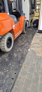 Wózek widłowy na podjeździe obok maszyny; pracownik naprawia podłogę. W tle widać elementy budowy.