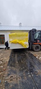 Transport dużego białego obiektu, przykrytego żółtą folią, na placu budowy obok budynku.