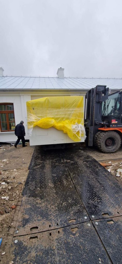 Transport dużego białego obiektu, przykrytego żółtą folią, na placu budowy obok budynku.