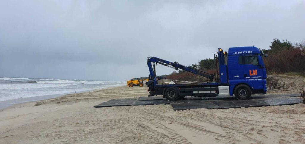 Ciężarówka na plaży z podnosnikiem, w tle wzburzone morze i chmurne niebo. Widać również drugi pojazd.