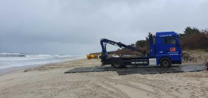 Ciężarówka na plaży z podnosnikiem, w tle wzburzone morze i chmurne niebo. Widać również drugi pojazd.