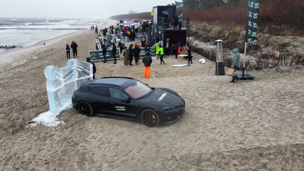 Na plaży stoi samochód, obok lodowy rzeźba i grupa ludzi w otoczeniu. Morze w tle, szare niebo.