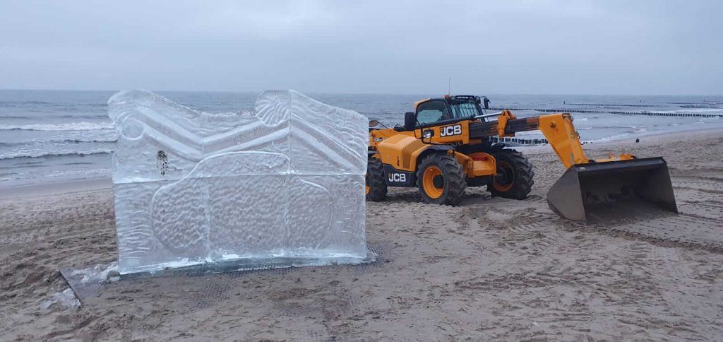 Na plaży stoi duża rzeźba lodowa obok żółtego ciągnika JCB, w tle widoczne fale morza.