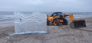 Na plaży stoi duża rzeźba lodowa obok żółtego ciągnika JCB, w tle widoczne fale morza.