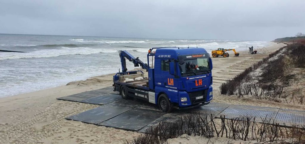 Niebieski samochód ciężarowy z platformą na plaży, obok pracująca maszyna oraz fale morskie w tle.