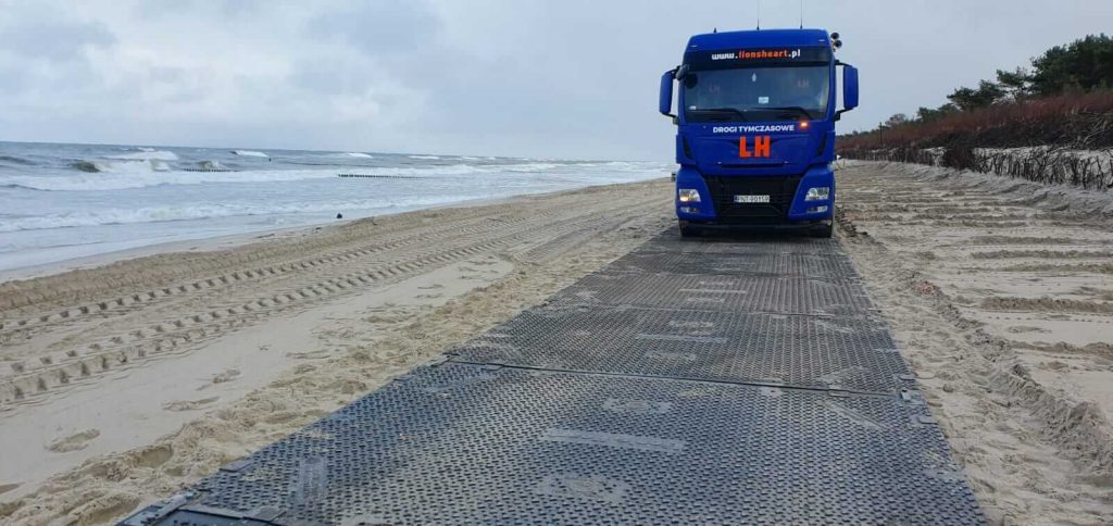 Niebieski ciężarówka stoi na plaży, na piasku leży metalowa nawierzchnia, w tle morze i fale.