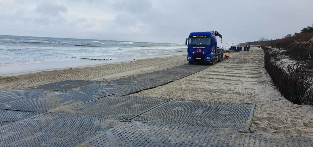 Na plaży widoczny jest niebieski ciężarówka, która przewozi materiały, obok leżą maty ochronne. W tle morze i chmury.