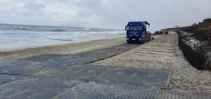 Na plaży widoczny jest niebieski ciężarówka, która przewozi materiały, obok leżą maty ochronne. W tle morze i chmury.