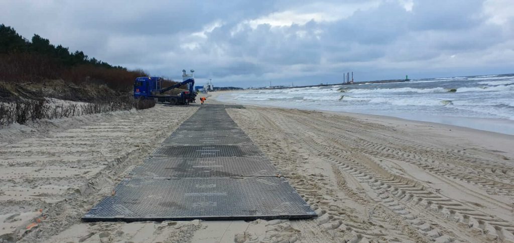 Na plaży widać umieszczone płyty, pojazd budowlany oraz fale morskie w tle. Niebo jest pochmurne.
