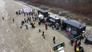 Ludzie na plaży w okolicy mobilnych stoisk, w tle las, część osób w kurtkach, lekki deszcz.