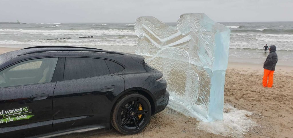 Samochód na plaży obok dużego bloku lodu, falujące morze w tle, deszczowa pogoda i osoba w stroju przeciwdeszczowym.