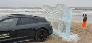 Samochód na plaży obok dużego bloku lodu, falujące morze w tle, deszczowa pogoda i osoba w stroju przeciwdeszczowym.