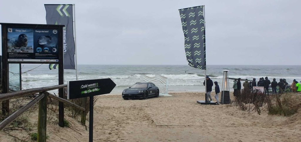 Na plaży stoi czarny samochód, obok flagi i tablica informacyjna. W tle widać morze i falujące fale.