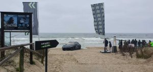 Na plaży stoi czarny samochód, obok flagi i tablica informacyjna. W tle widać morze i falujące fale.