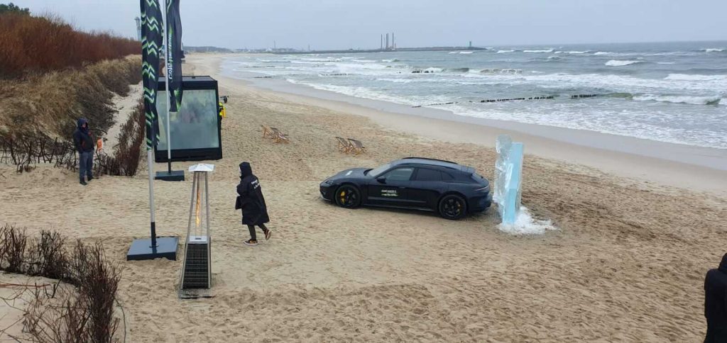 Na plaży widać czarny samochód, rzeźbę z lodu oraz osoby w strojach zewnętrznych. W tle morze i molo.