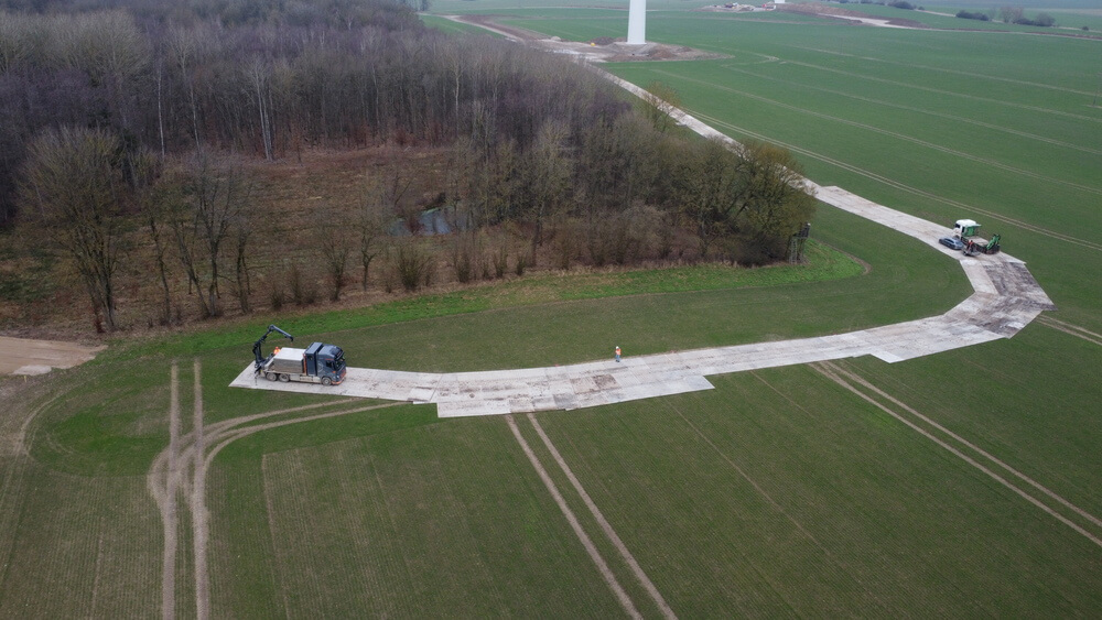 Widok z lotu ptaka na pole, na którym znajdują się maszyny budowlane i nowa nawierzchnia drogowa. Tło to las.