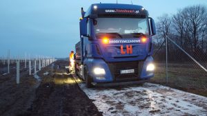 Ciężarówka z napisem LH stoi na drodze, obok pracownik w kamizelce odblaskowej wykonuje prace budowlane.