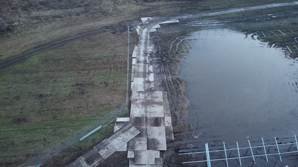 Brudna ścieżka prowadząca przez błotnisty teren obok stawu, z widocznymi płytami betonowymi.