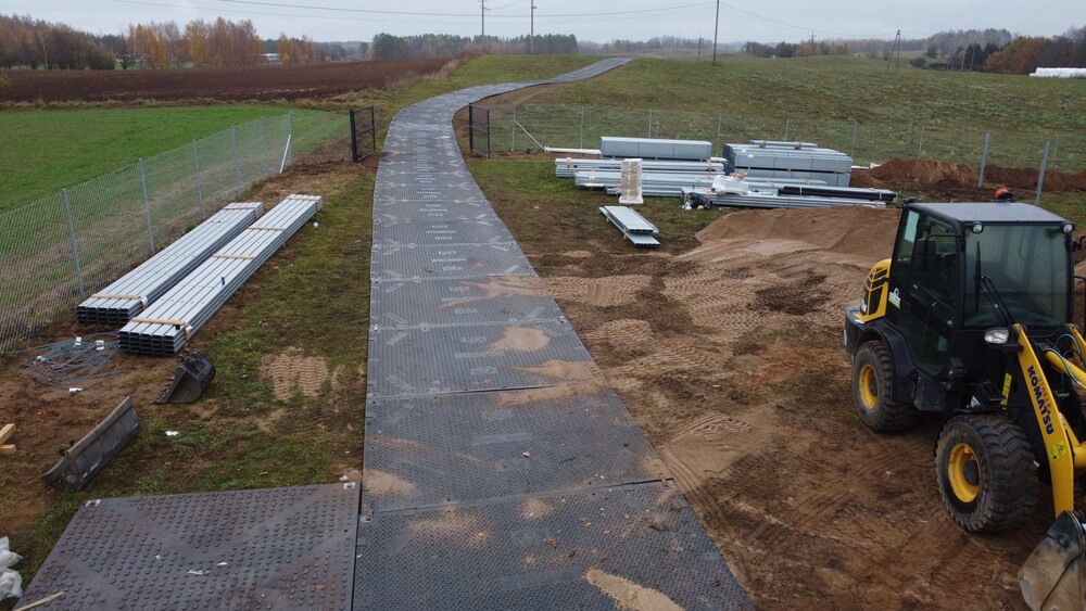 Widok na plac budowy z drogą utwardzoną, materiałami budowlanymi i maszyną budowlanych. Tło to pola i drzewa.