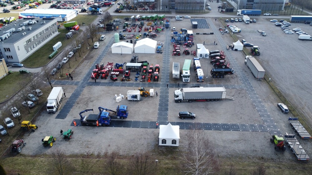 Widok z góry na plac pełen maszyn rolniczych, przyczep, ciężarówek i namiotów, otoczony samochodami w tle.