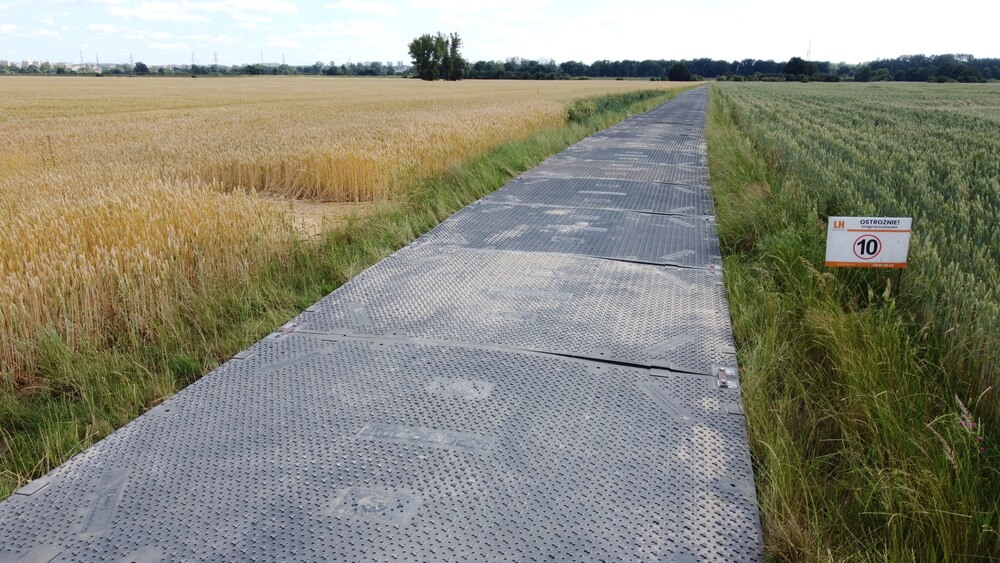 Droga z płyt betonowych prowadzi przez pola zbóż i trawy, z tablicą ostrzegawczą z ograniczeniem prędkości.