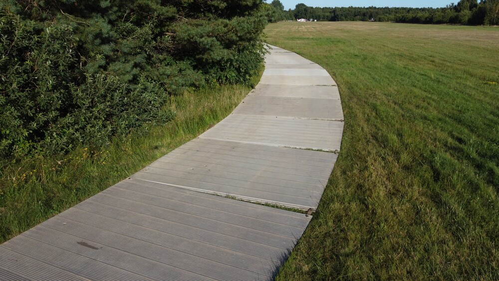 Ścieżka rowerowa wykonana z desek biegnąca przez zieloną łąkę, otoczona drzewami.