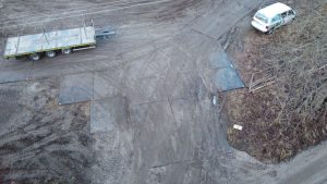 Widzimy fragment drogi z wyrównanym, błotnistym podłożem, obok zaparkowany samochód i przyczepę.