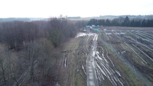 Widok na błotnistą drogę w lesie, obok pola z kłami oraz budynkami w tle. Deszczowa, ponura atmosfera.