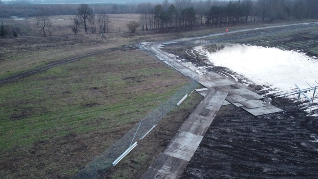 Widok na teren budowy z zalaną powierzchnią, błotem i fragmentami ogrodzenia. W tle drzewa, a w oddali otwarta przestrzeń.