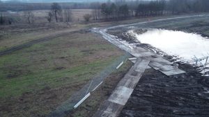 Widok na teren budowy z zalaną powierzchnią, błotem i fragmentami ogrodzenia. W tle drzewa, a w oddali otwarta przestrzeń.