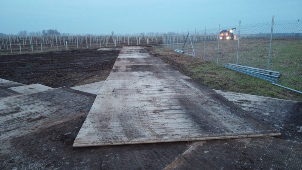 Fotografia przedstawia plac budowy z ułożonymi drewnianymi deskami oraz ogrodzeniem, w tle widoczny traktorek i pola.