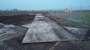 Fotografia przedstawia plac budowy z ułożonymi drewnianymi deskami oraz ogrodzeniem, w tle widoczny traktorek i pola.