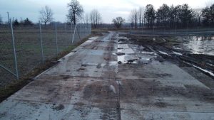 Fragment terenu z ułożonymi płytami betonowymi, otoczony ogrodzeniem, na tle barren krajobrazu i drzew.