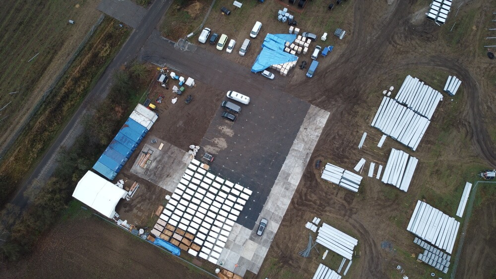 Widok z góry na plac z rozłożonymi materiałami budowlanymi i pojazdami. W tle pola, w dolnej części płyta betonowa.