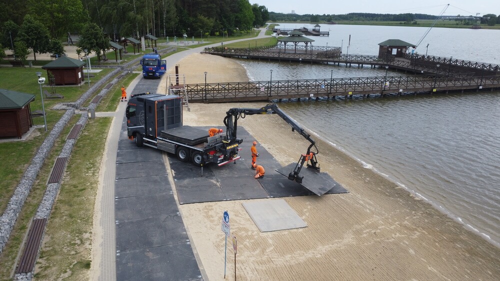 Ciężarówka z podnośnikiem na plaży, pracownicy w pomarańczowych ubraniach, nad wodą i w pobliżu molo.