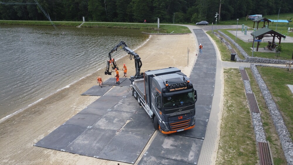 Ciężarówka z dźwigiem na plaży, obok ekipy pracującej, w tle woda i tereny rekreacyjne.